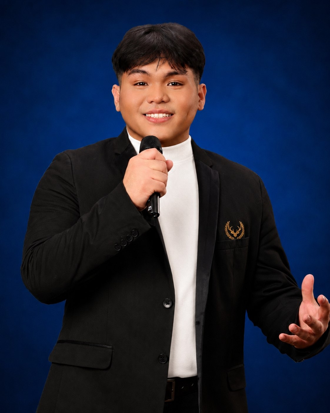 Ralskies holding a microphone against a blue background, wearing a black blazer with a gold antler emblem